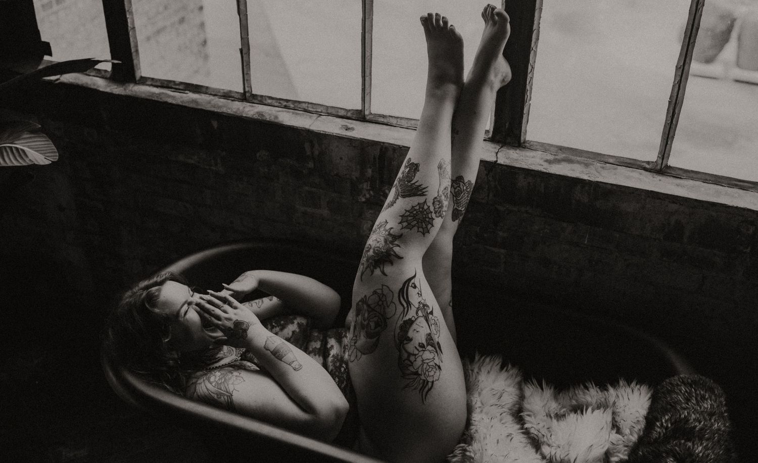 Figure in black lace bodysuit poses near window with sheer white curtains in moody boudoir photography session.