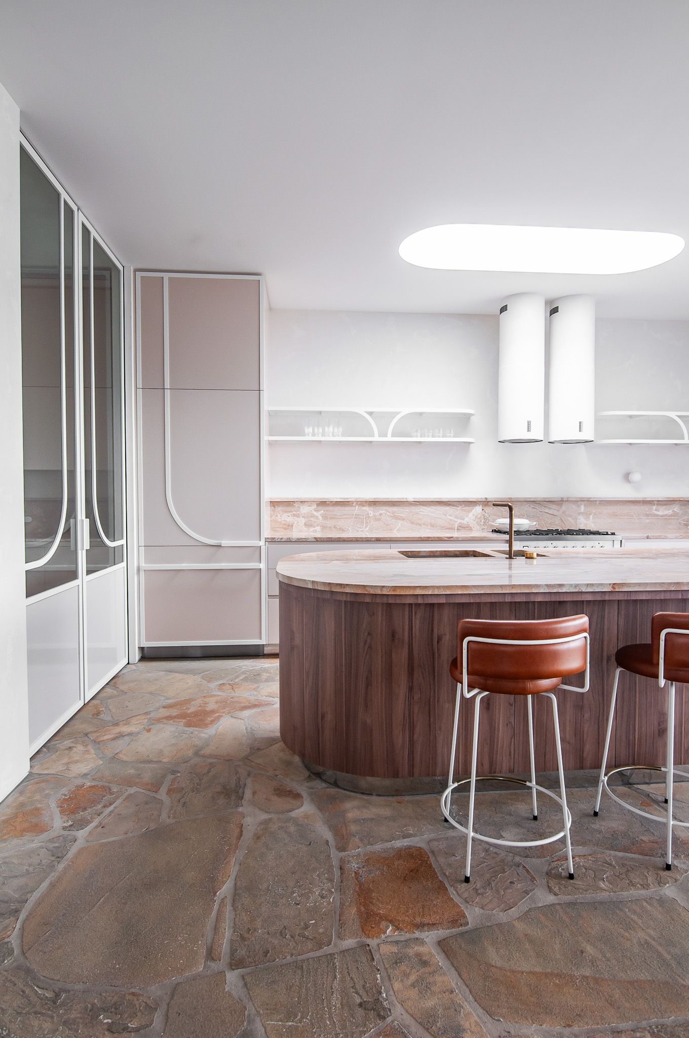 Modern kitchen featuring curved wooden island, leather barstools, and stone flooring with pink undertones.
