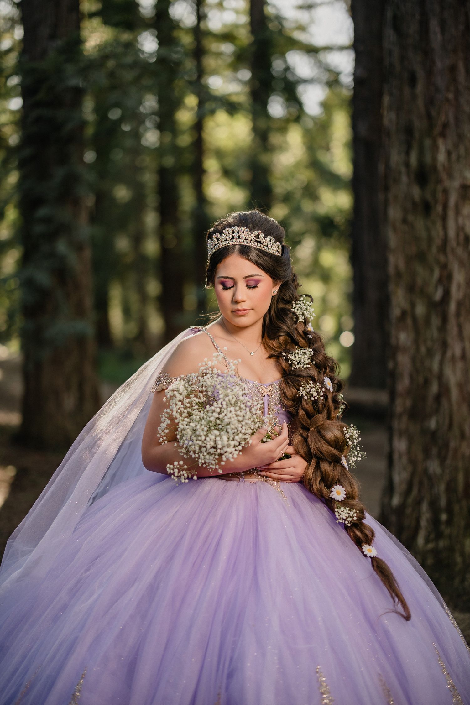 A person in a lavender quinceañera dress with a crown and long curled hair poses in a forest setting.