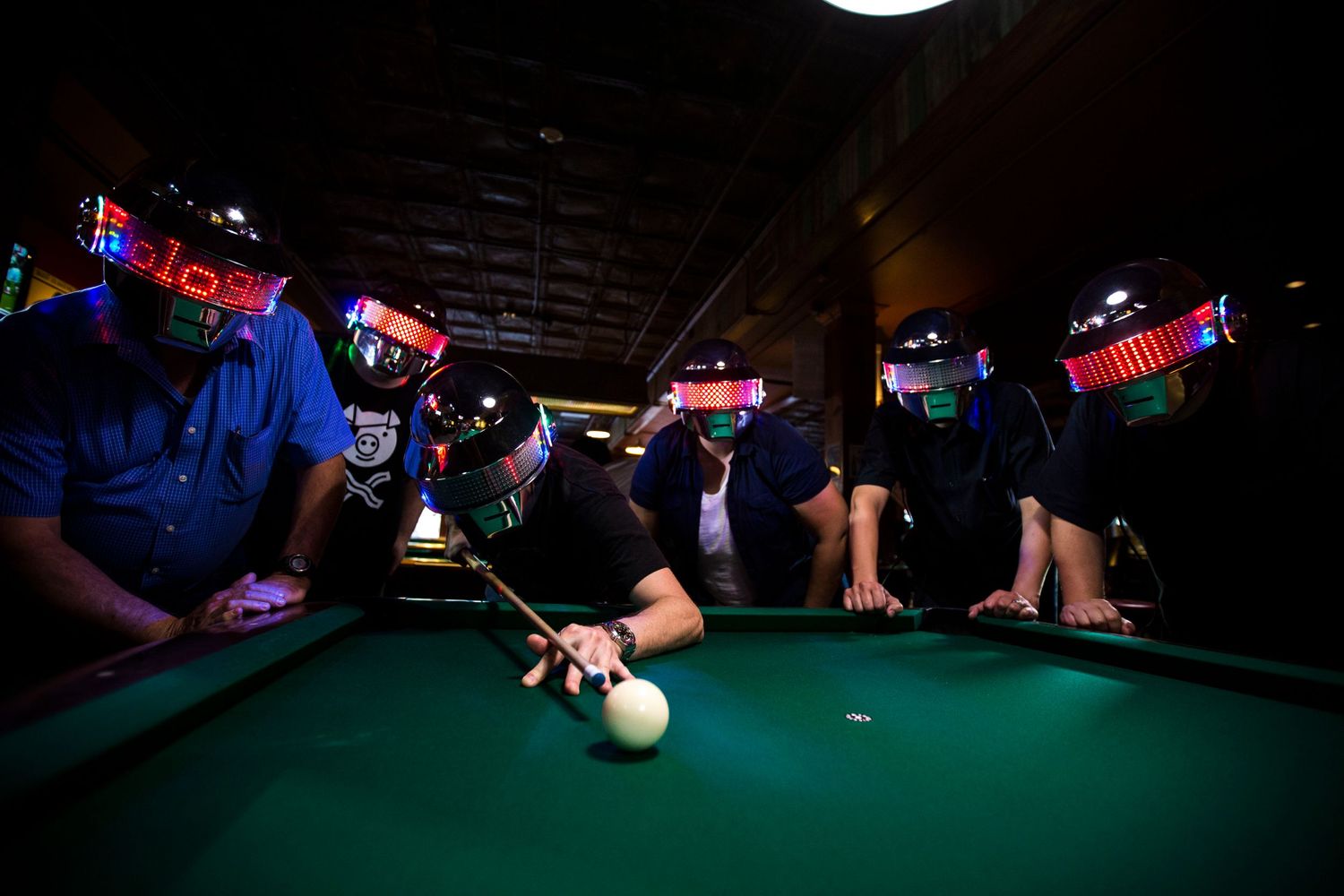 People play pool at a dimly lit bar with neon lights glowing overhead while someone takes a shot at the table.