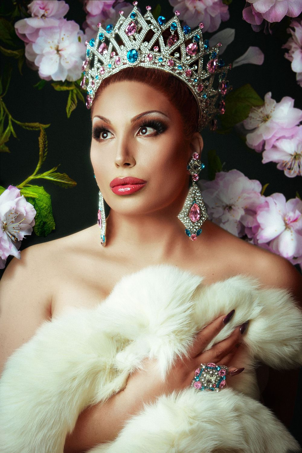 Glamorous portrait with sparkling crown, jeweled earrings and white fur against floral backdrop.