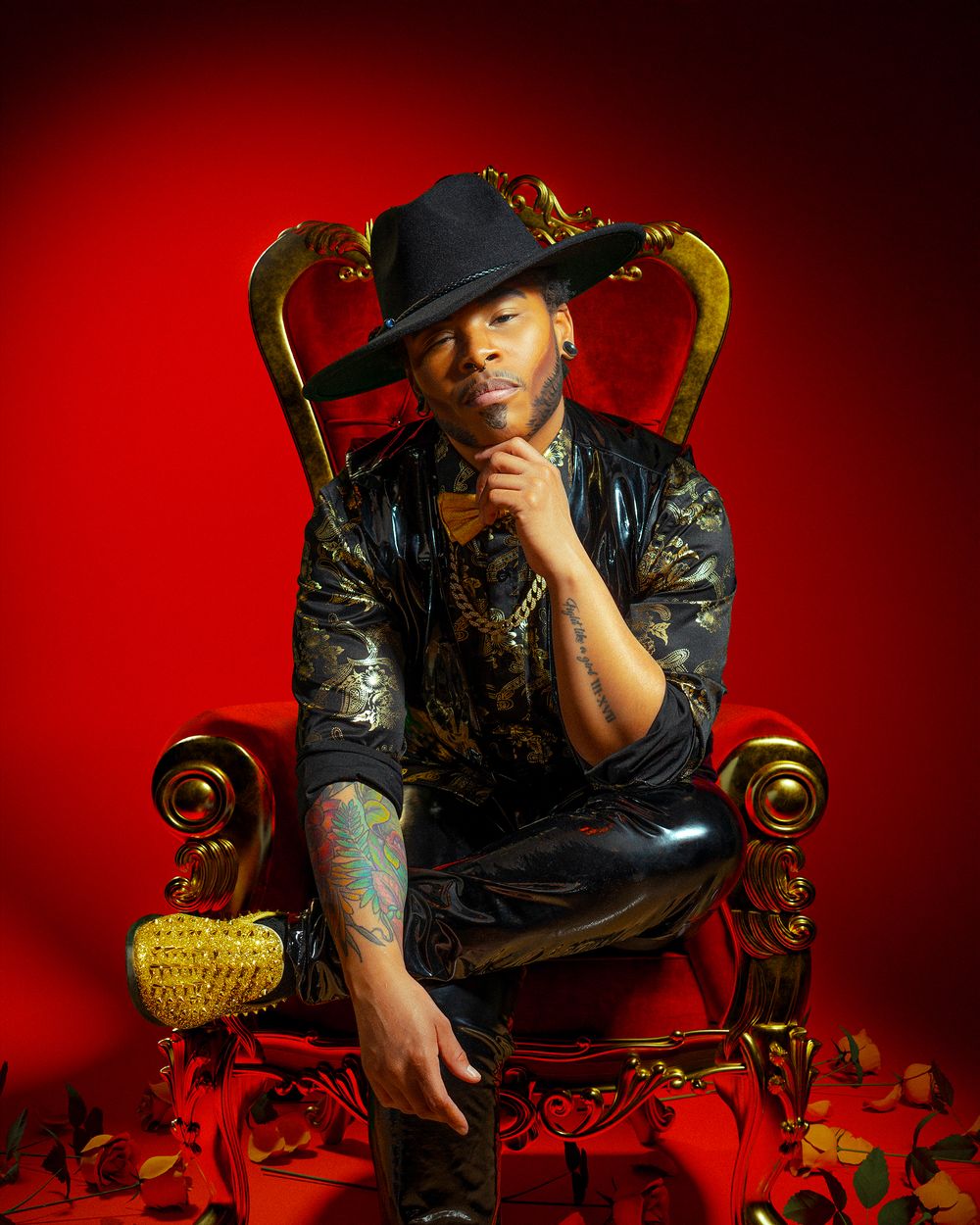 Stylish figure in black attire and hat poses on ornate throne against red backdrop.