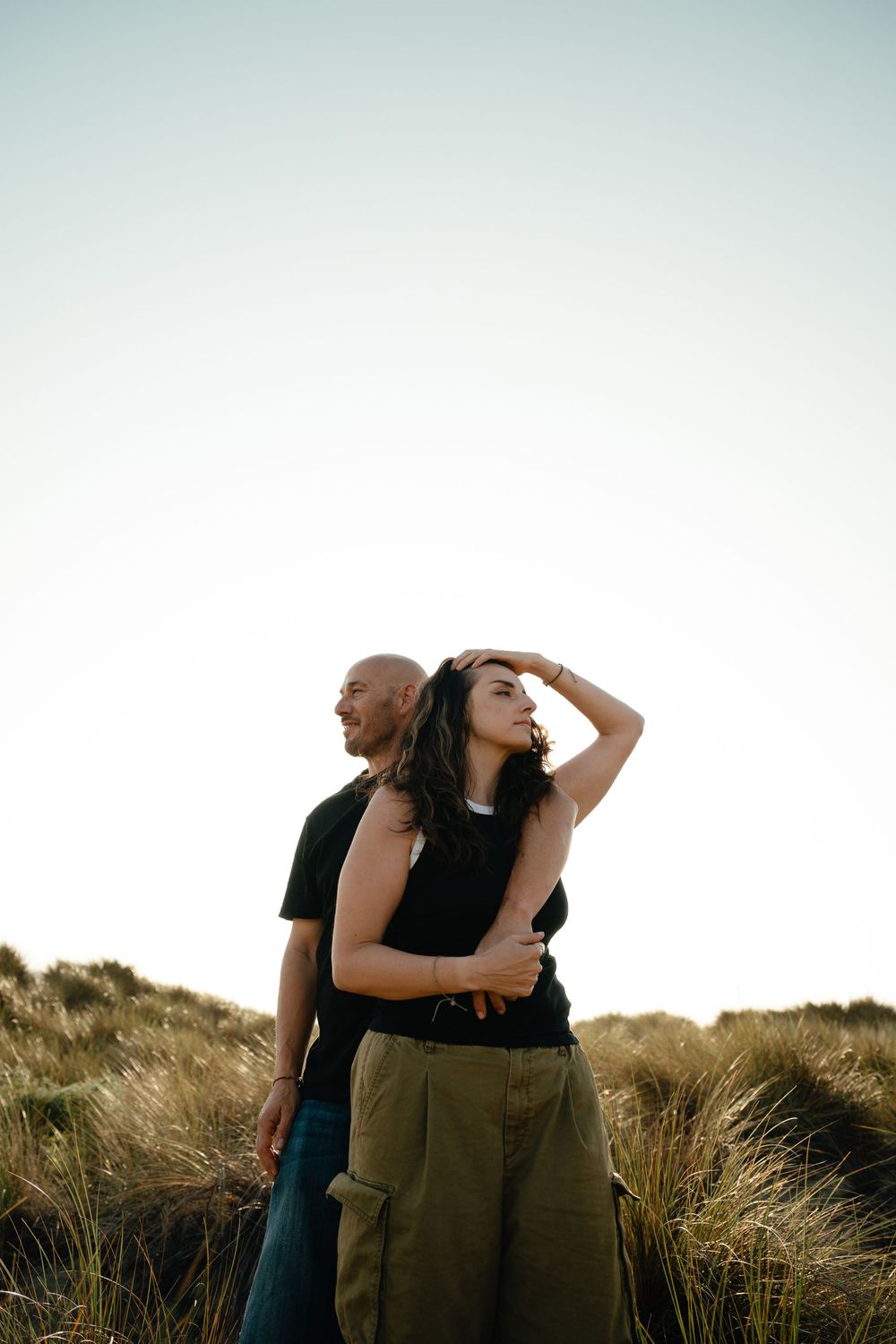 Couple standing in tall grass against bright sky during golden hour in romantic outdoor setting.