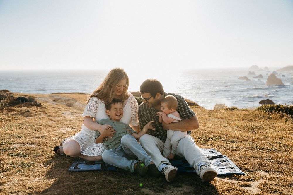 Family sits together on coastal cliff during golden hour while looking at something on a device.