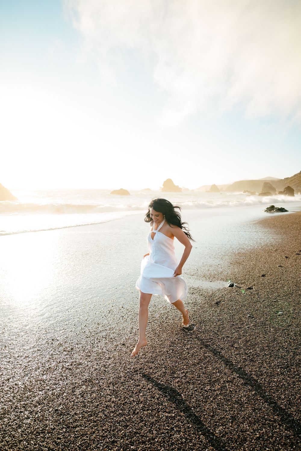 Someone in a flowing white dress runs joyfully along a rocky beach at sunset with coastal cliffs in the background.