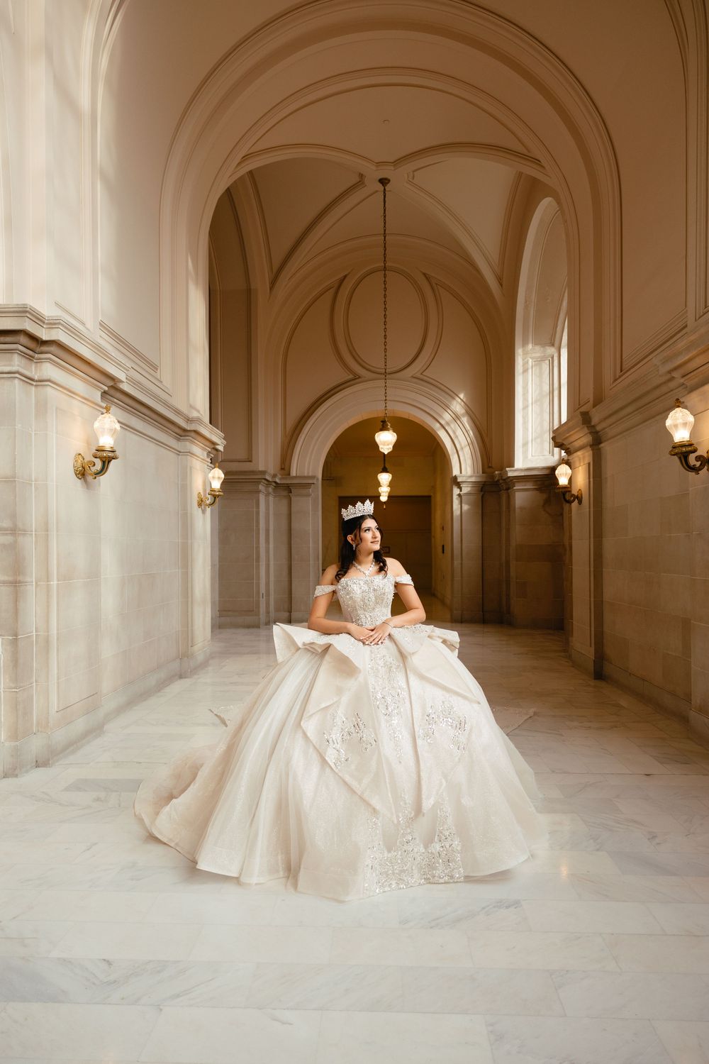 A person in an ornate white ball gown stands in an elegant hallway with arched ceilings and wall sconces.