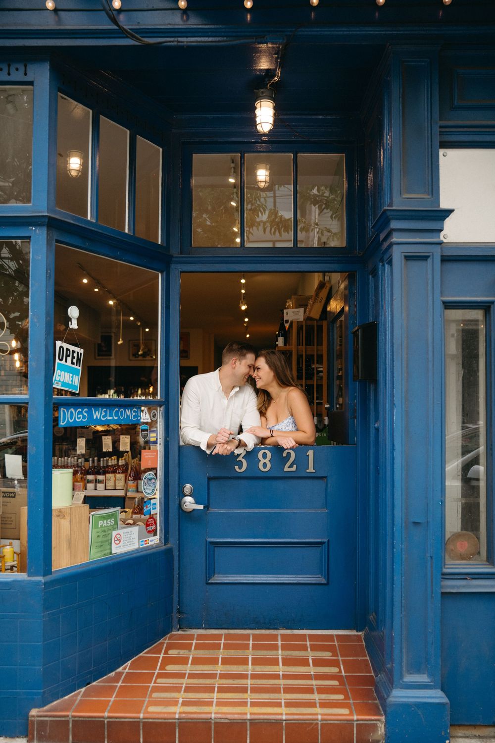Couple sharing a romantic moment in the doorway of a blue storefront numbered 3821 with warm interior lighting.