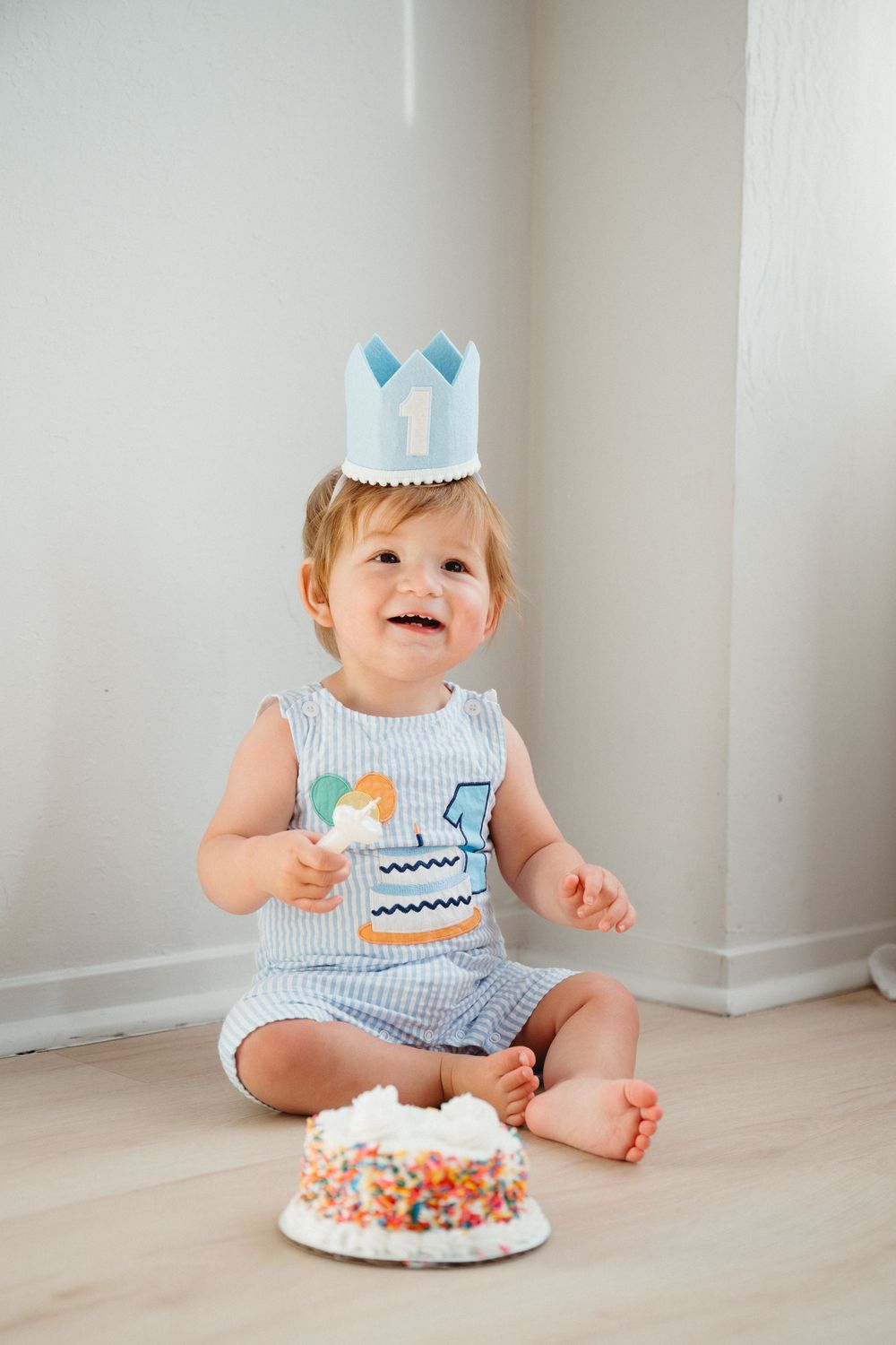 Baby in blue and white striped outfit sits on floor with birthday cake and smiling during first birthday celebration.