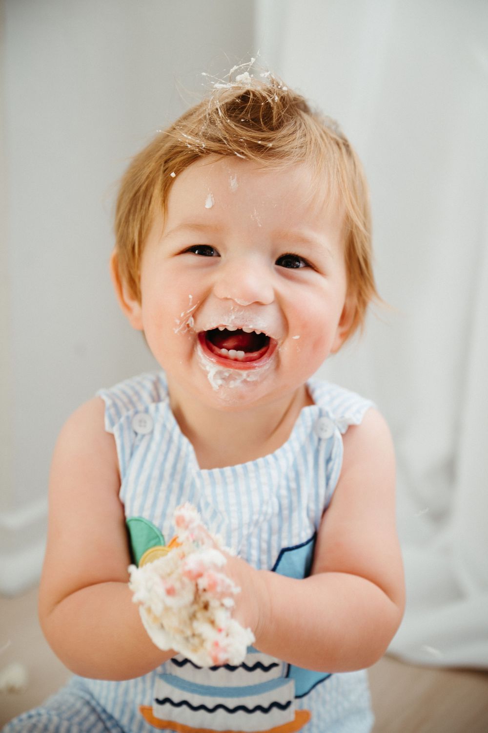 A happy toddler in a striped outfit enjoys a messy cake smash celebration with frosting on their face.