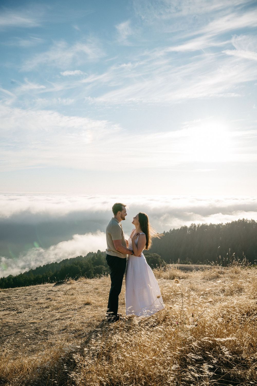 Couple embracing on a golden hilltop above the clouds at sunset, with misty mountains and blue sky behind them.