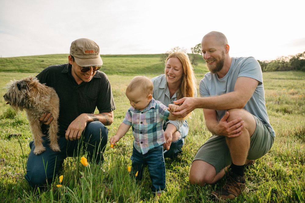 Family with baby and dog enjoying a sunny day in a green meadow, crouching together and smiling outdoors.