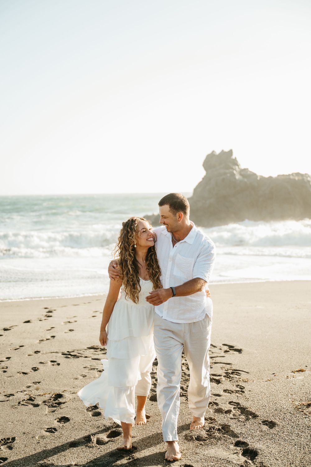 Happy couple in white outfits walking barefoot on a sunny beach, embracing and smiling at each other.