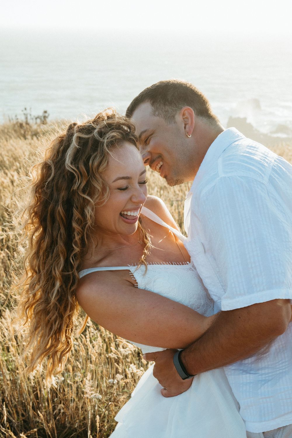 A joyful couple embraces and laughs together on a sunlit coastal bluff with golden grass and ocean views.