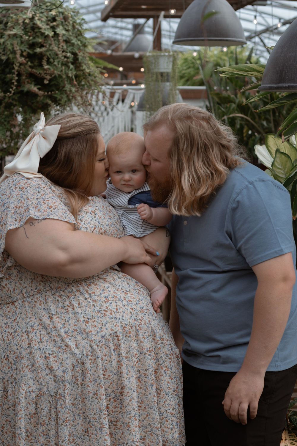 A family shares a loving moment in a greenhouse setting with hanging plants.