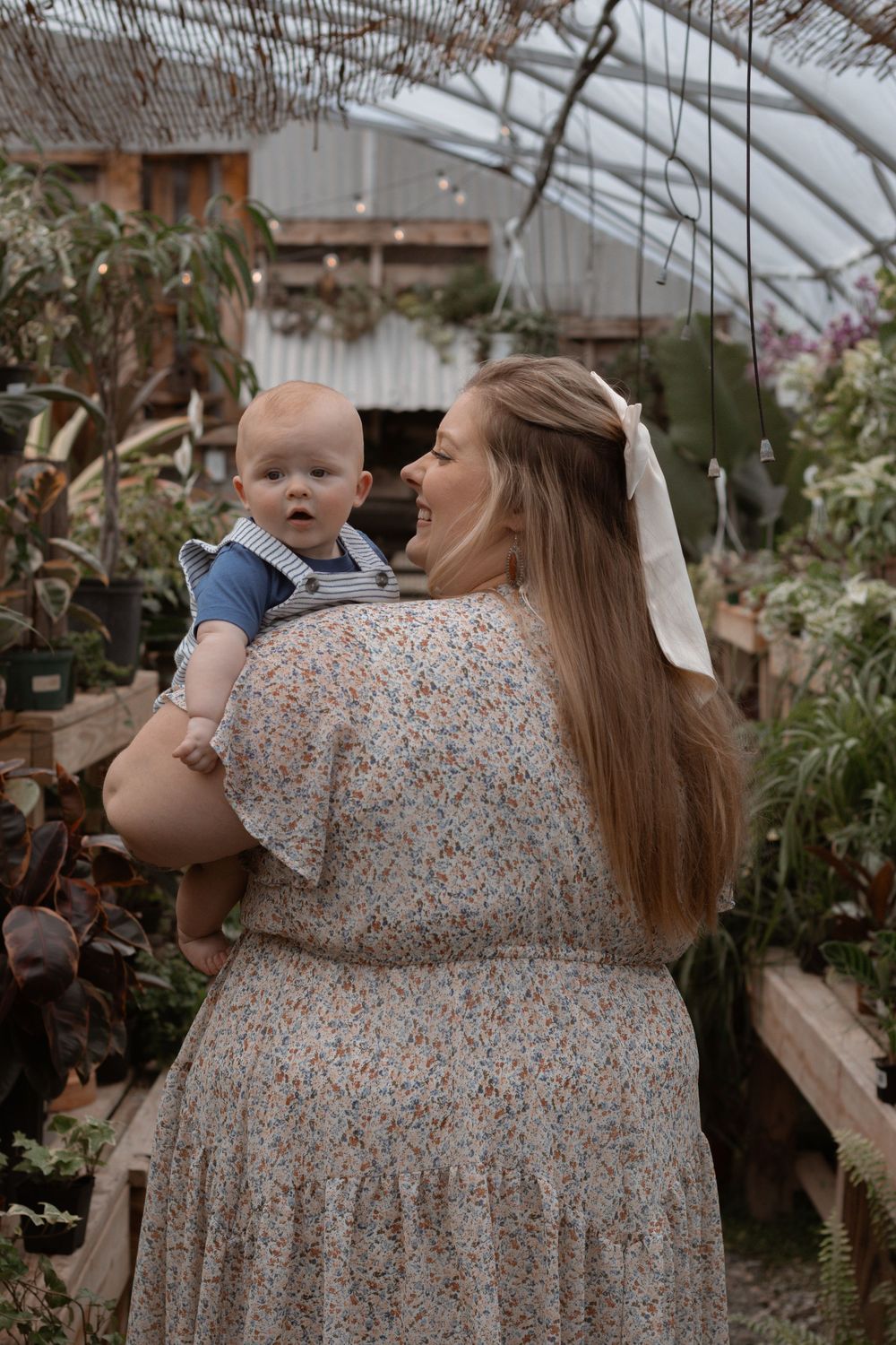 A tender moment between parent and baby inside a greenhouse filled with plants and flowers.