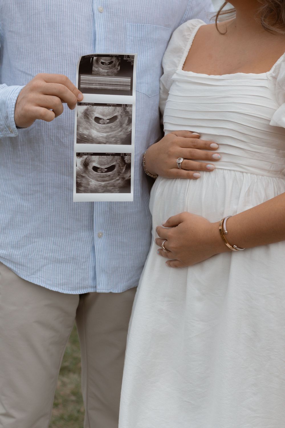 A couple holds ultrasound images while showcasing a white maternity dress and baby bump in an outdoor setting.