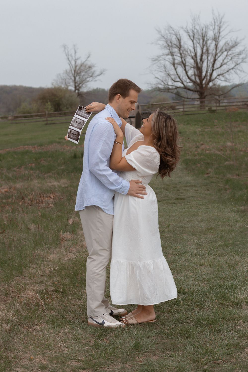 A romantic couple embraces in a field while holding ultrasound photos to announce their pregnancy.