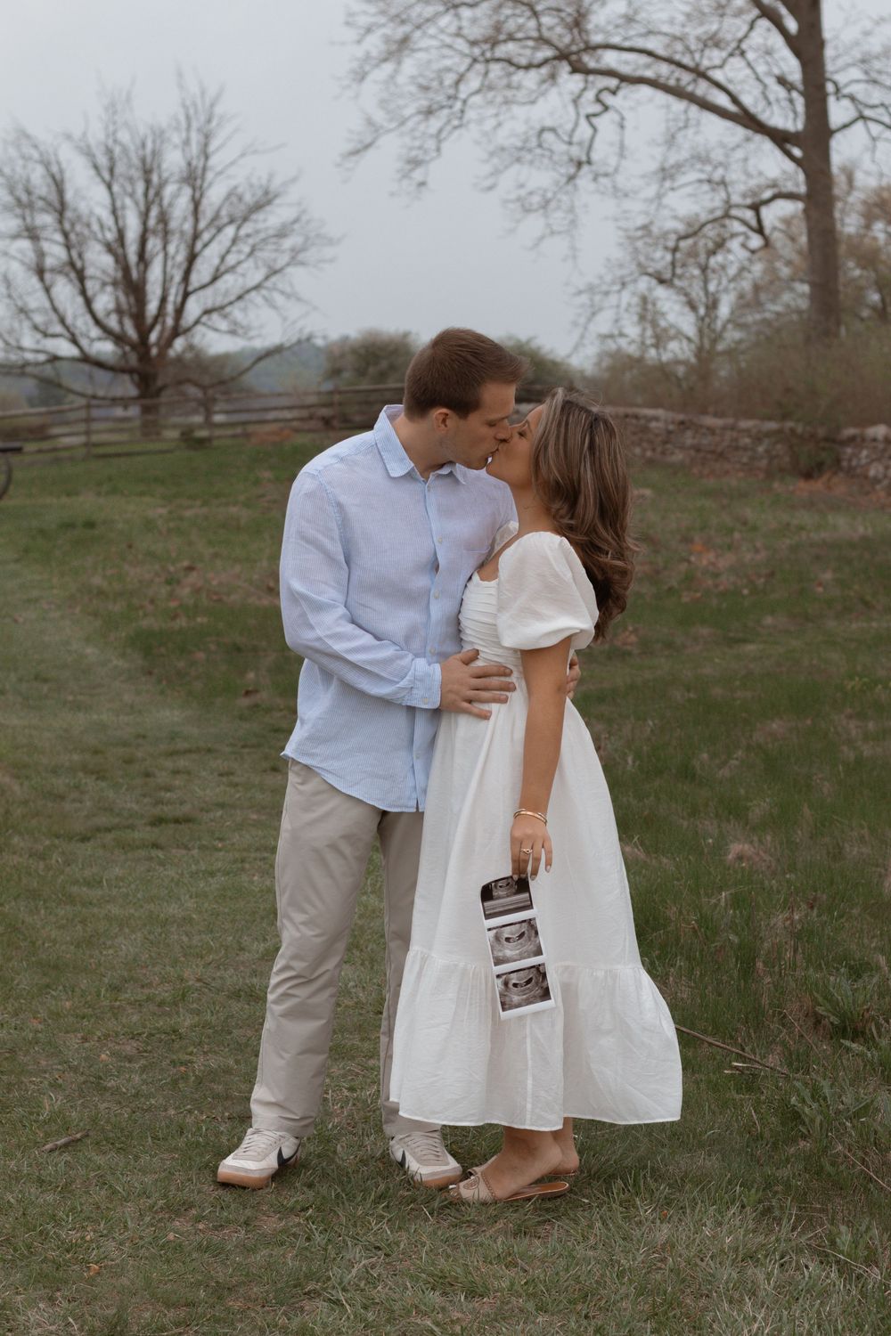 A tender moment between expecting parents sharing a kiss while holding ultrasound photos in a rustic field.