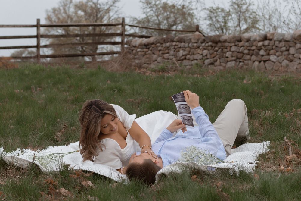 Expectant couple shares an intimate moment while viewing ultrasound photos on a blanket outdoors.