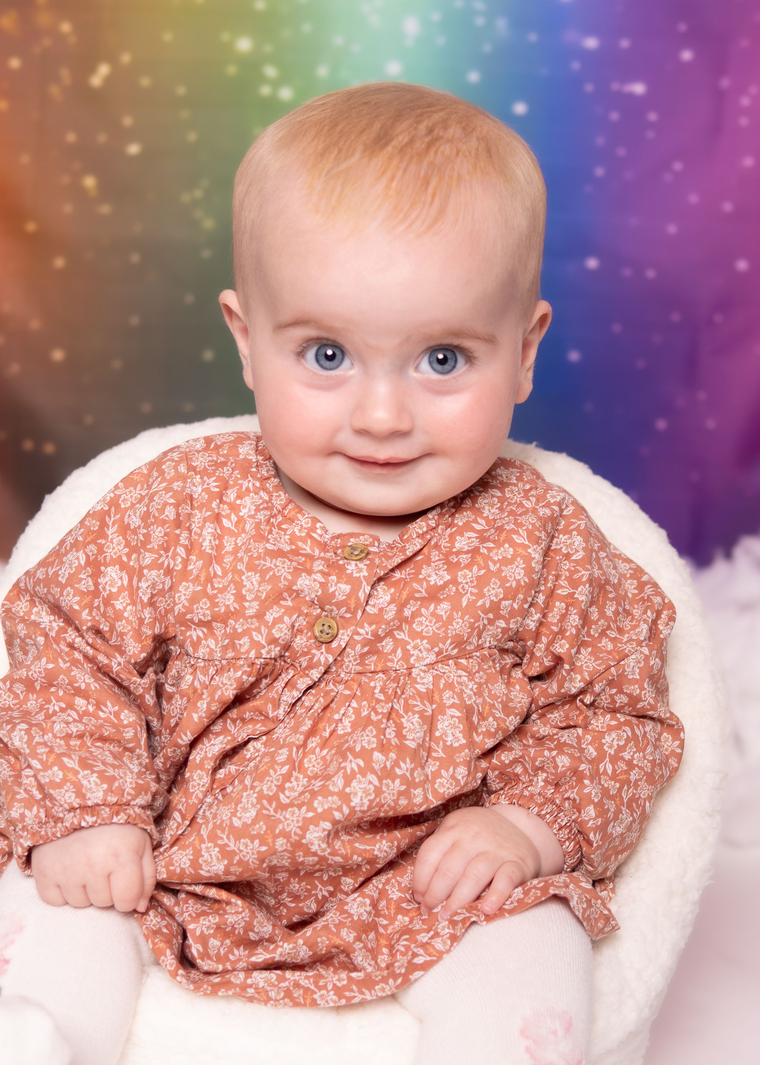 Baby wearing floral print outfit smiles while lying on white blanket against colorful rainbow background.