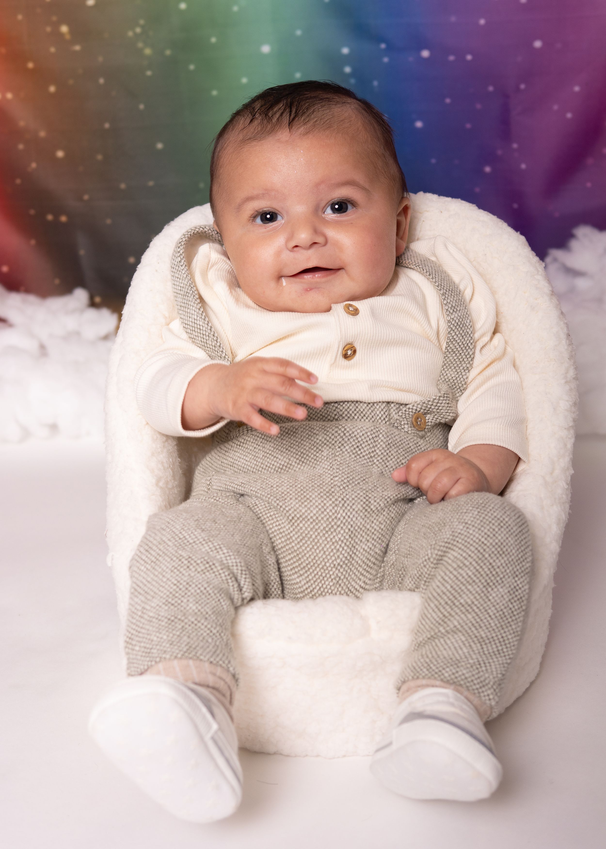 Baby in cream colored outfit rests on white cushion against rainbow colored backdrop.