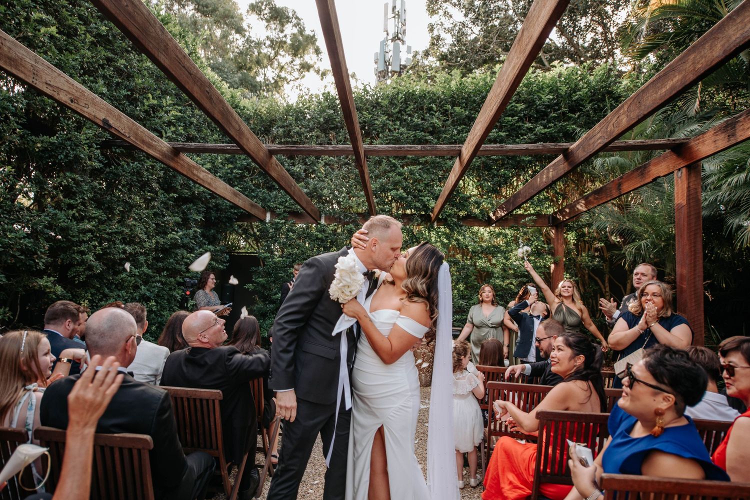 Wedding ceremony with guests celebrating under a rustic wooden pergola decorated with greenery.