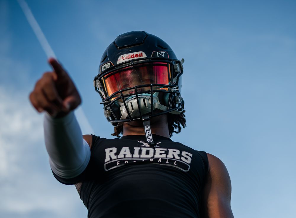 Football player in Raiders jersey and helmet pointing forward against a blue sky backdrop.