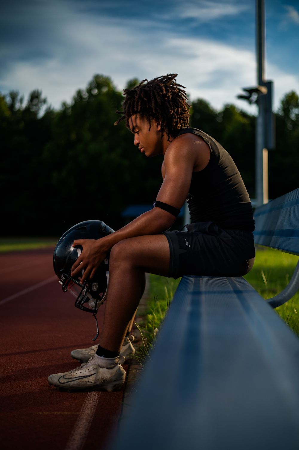 Young football player with dreadlocks sits on bleachers holding helmet, wearing cleats and athletic wear at dusk.