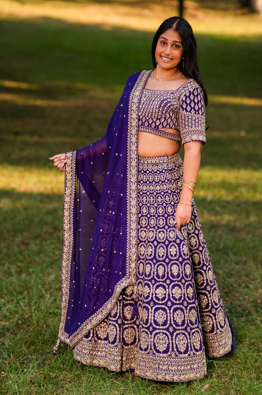 Young woman in ornate purple and gold lehenga choli with dupatta, posing outdoors on green grass.