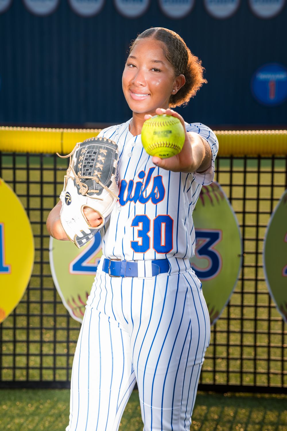 Softball player #30 in pinstripe uniform poses with glove and yellow ball on field.