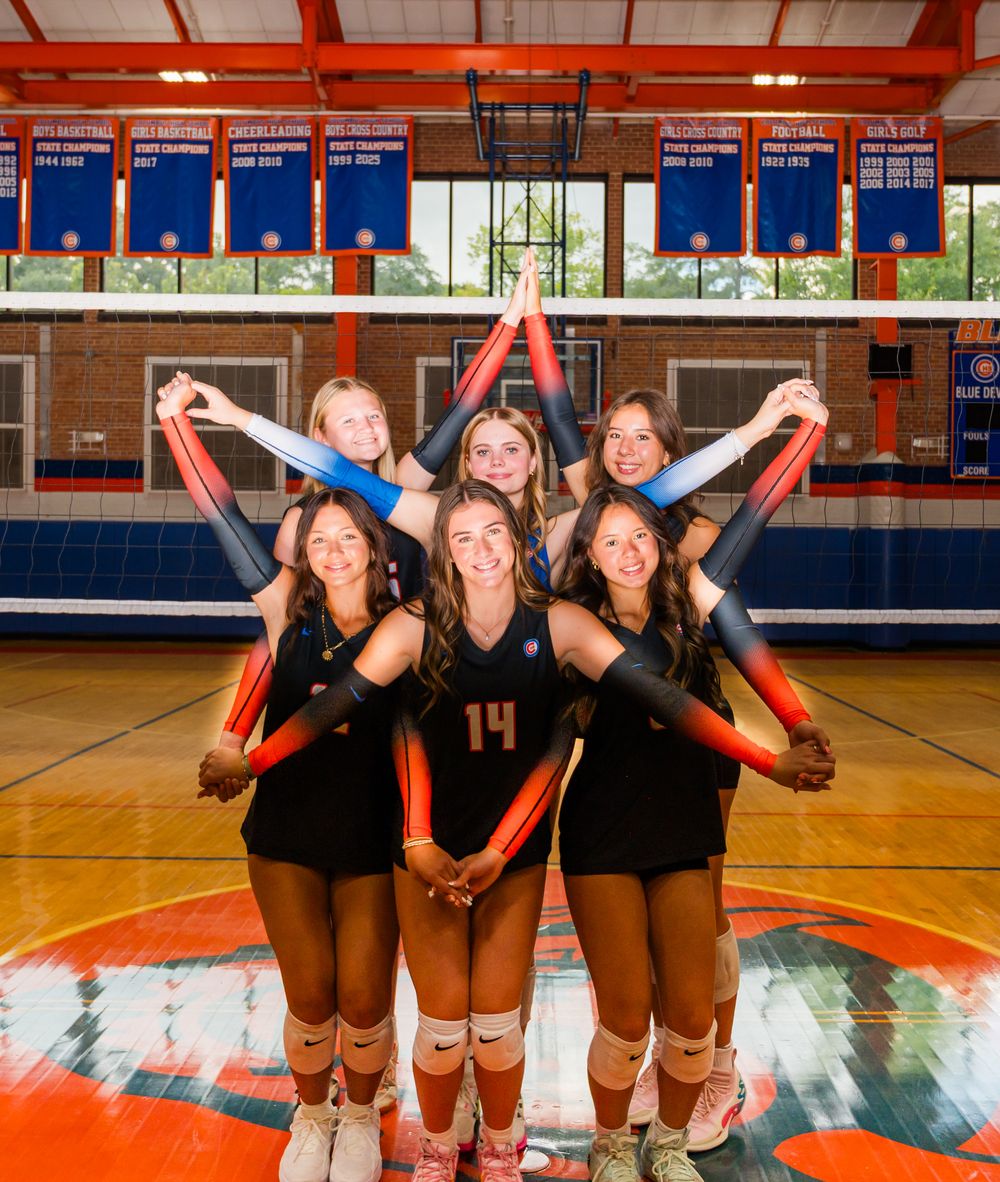Seven female volleyball players pose in a gym holding red and blue foam sticks, wearing black uniforms with championship banners above.