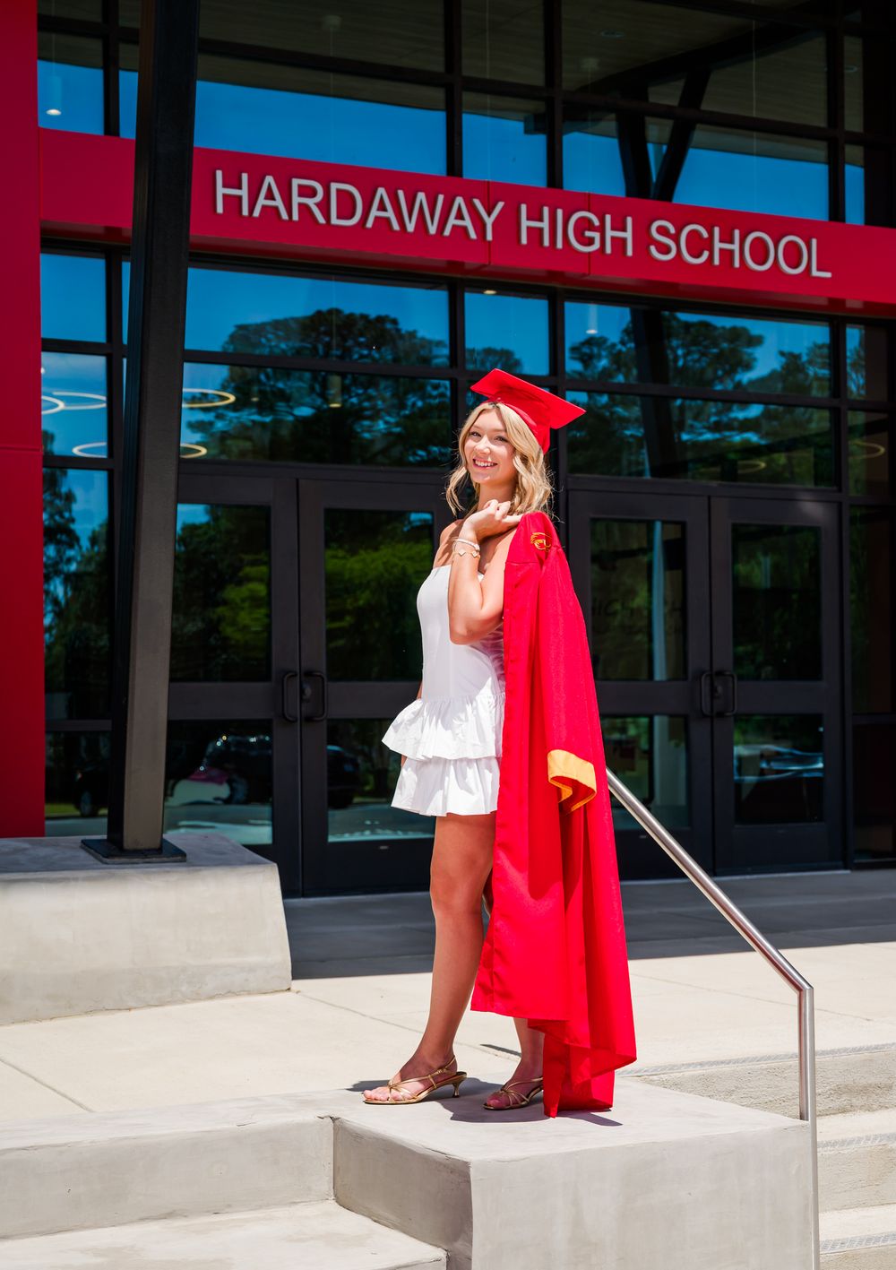A graduate in red cap and gown sits reflectively on concrete steps outside Hardaway High School.