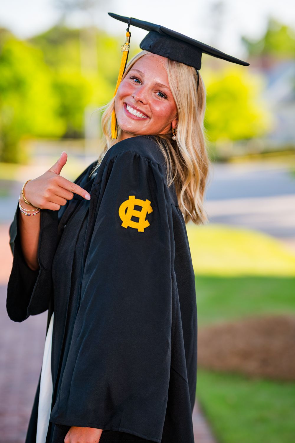 Smiling blonde graduate in black cap and gown pointing to gold HC logo on sleeve outdoors.