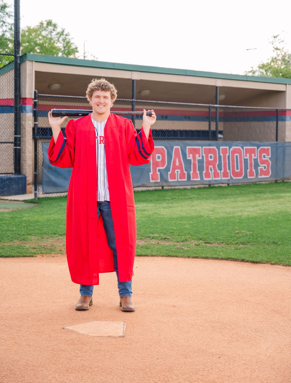 Graduate in red cap and gown stands on baseball field near Patriots dugout during outdoor ceremony.