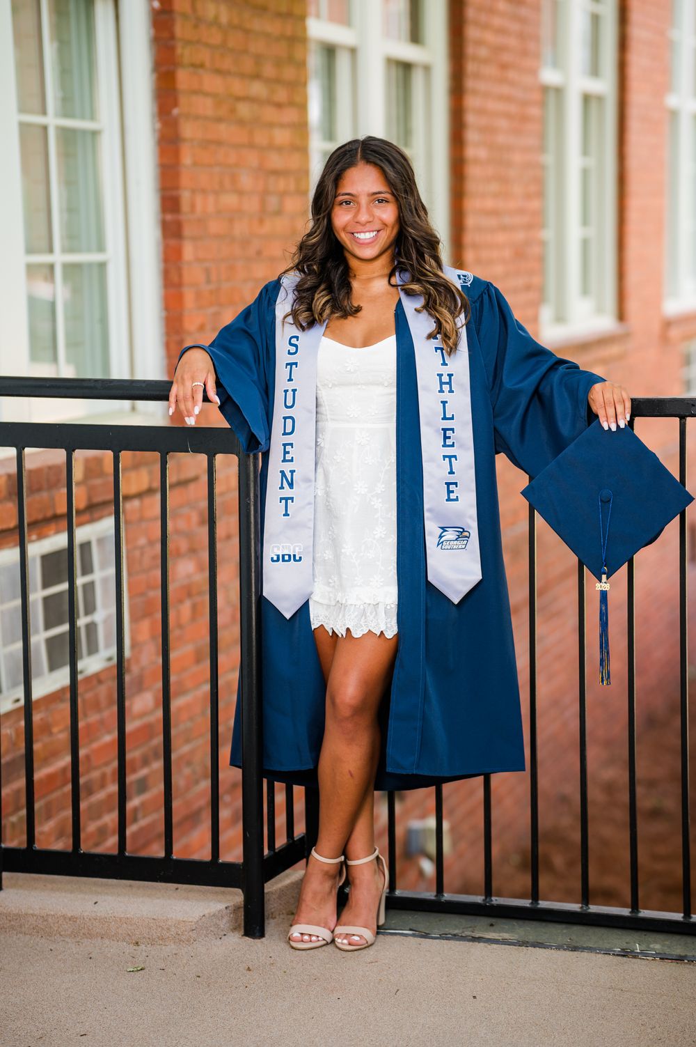 Smiling graduate in navy blue cap and gown with student athlete sash poses outdoors at brick building.