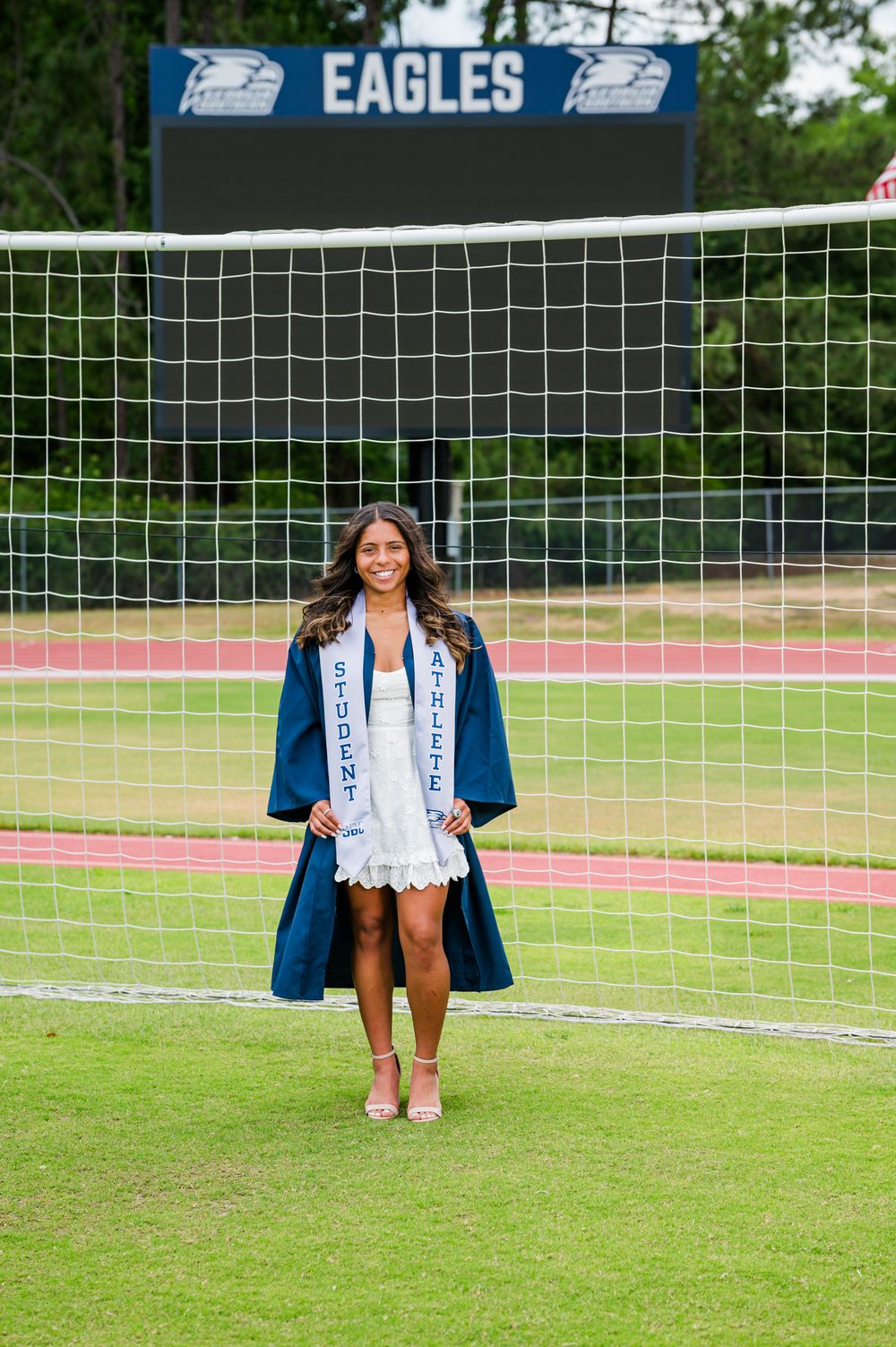 A smiling female student-athlete in navy graduation gown and white dress poses proudly in front of a soccer goal at Eagles stadium.
