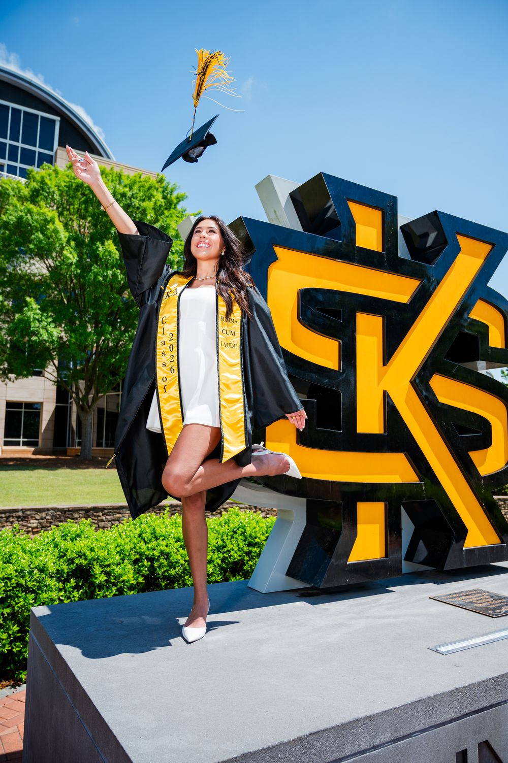 A joyful graduate in black and gold regalia poses triumphantly in front of Kennesaw State University's KSU sign.