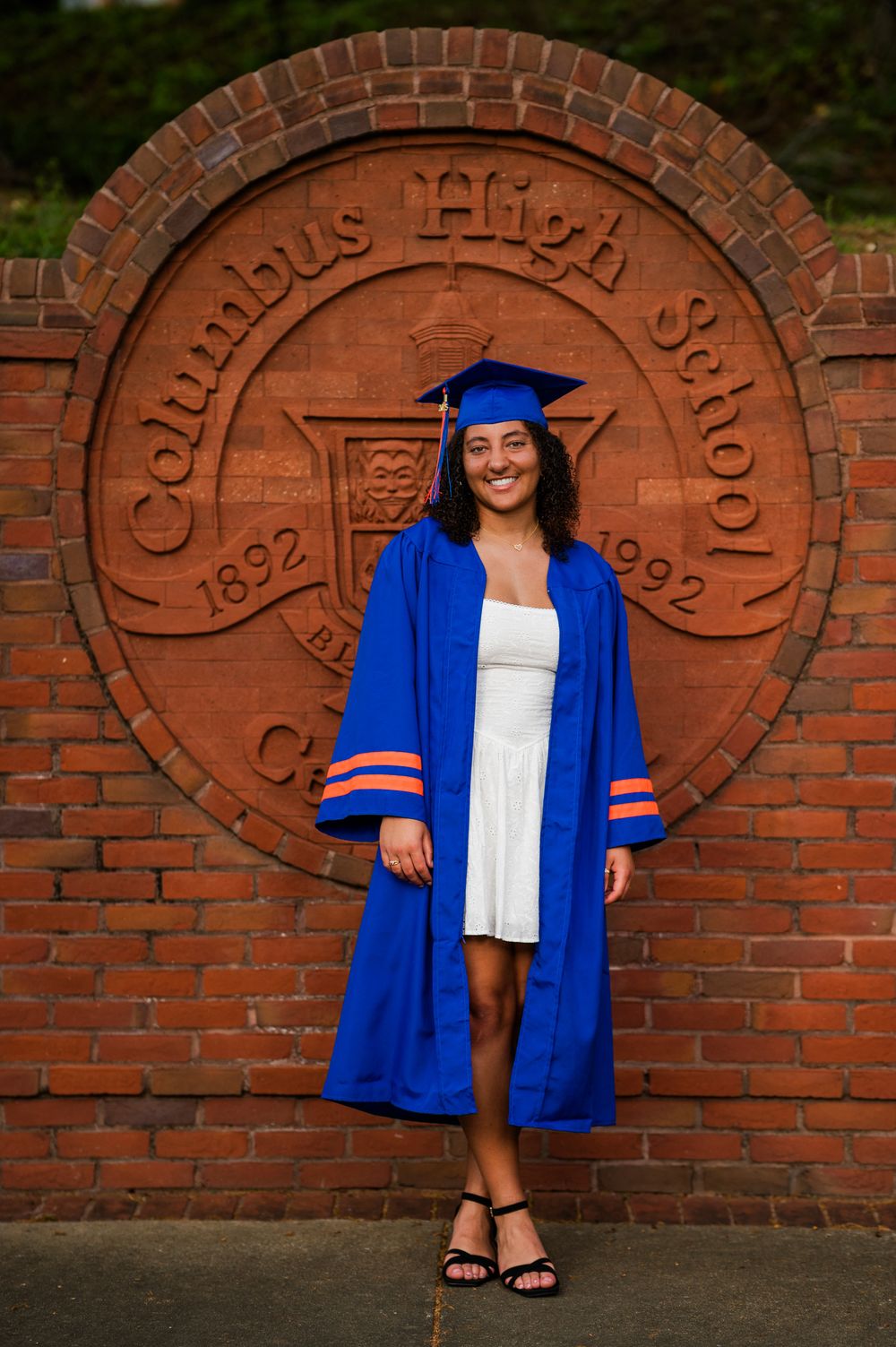 Graduate in royal blue cap and gown stands in front of brick wall with carved school seal at Columbus High School.