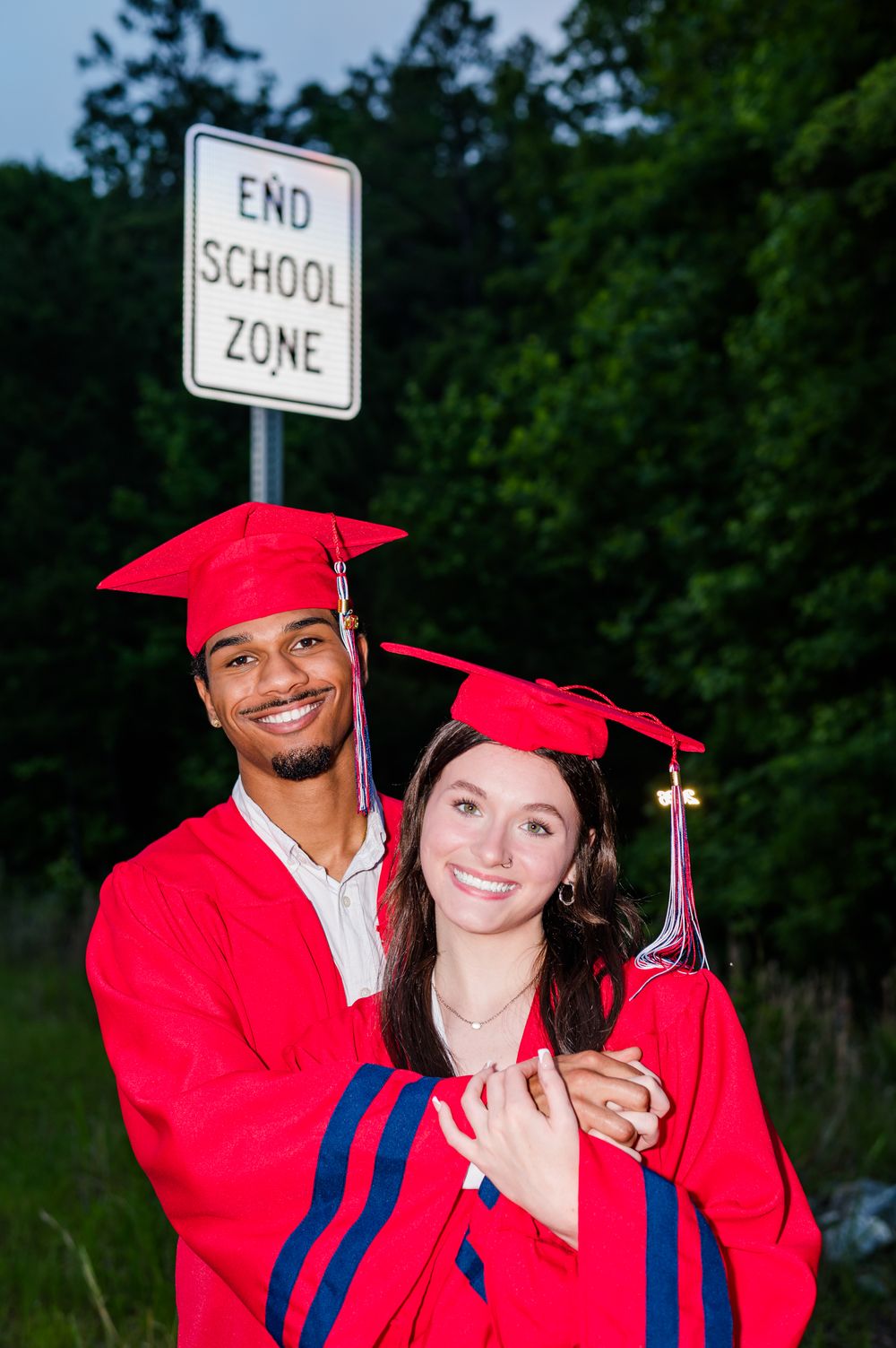 Two graduates in red caps and gowns smile in front of an End School Zone sign, celebrating their graduation.