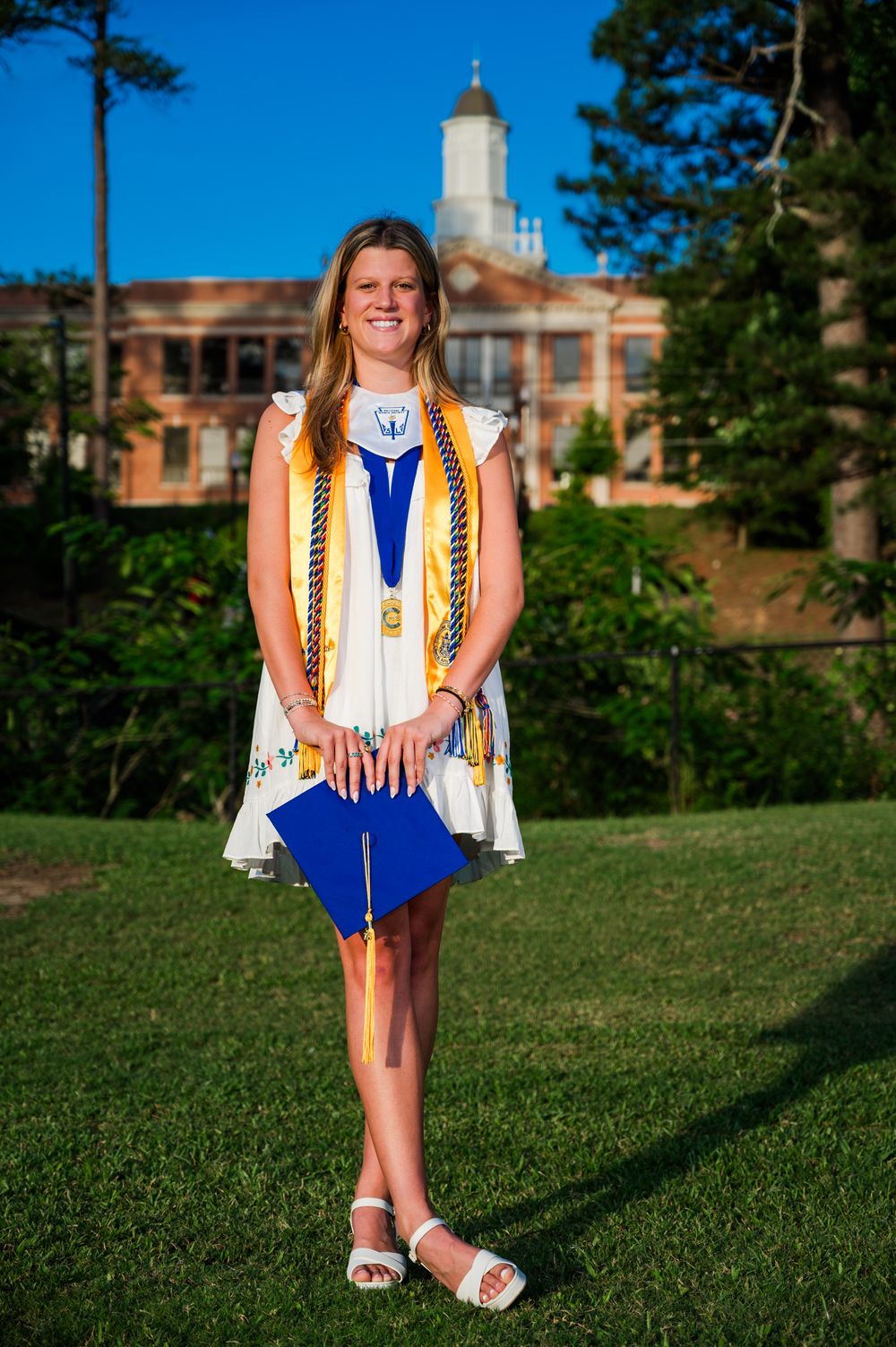 A person in a yellow striped vest and blue dress stands on green grass with brick buildings in the background.