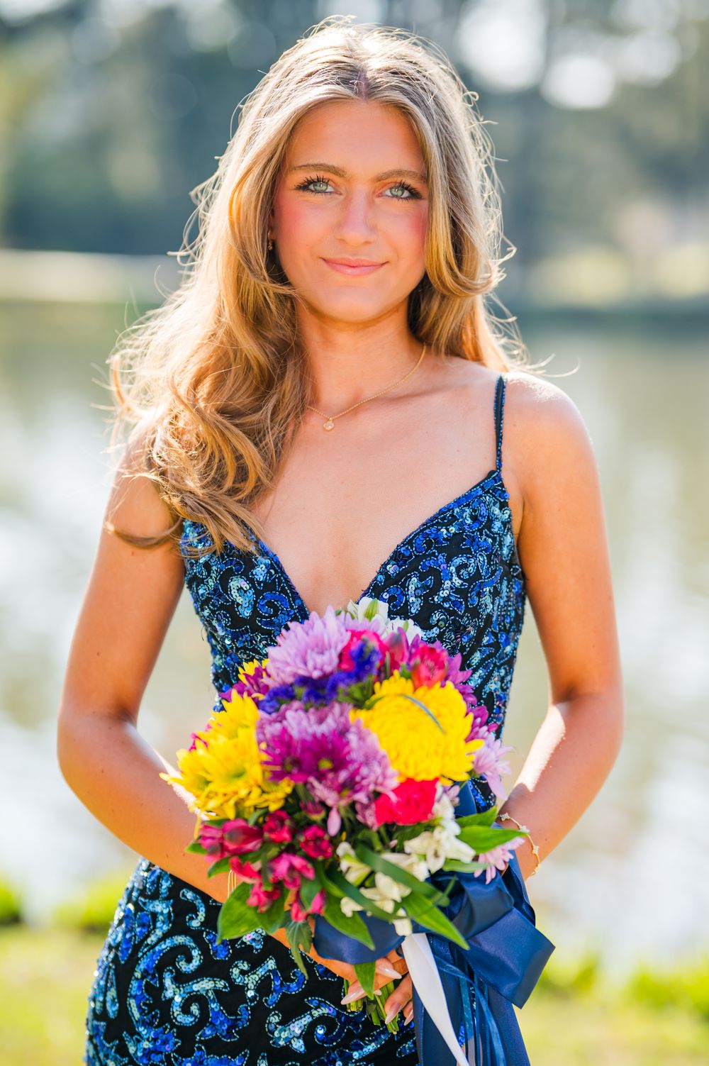 Young woman in blue sequin dress holding colorful flower bouquet, smiling outdoors near water.