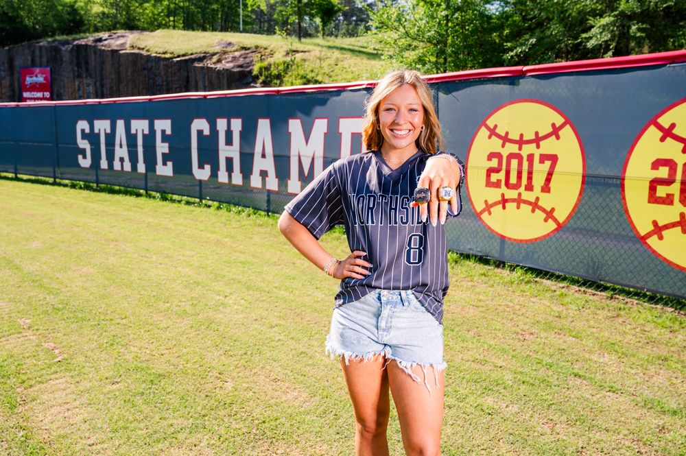 Young female athlete in softball jersey poses proudly at a 2017 State Championship field banner on a sunny day.