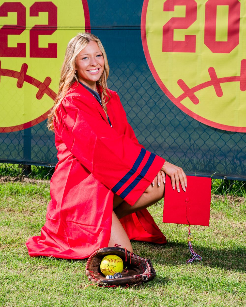 Senior girl in red graduation gown poses with softball glove and ball in front of Class of 2022 banner.