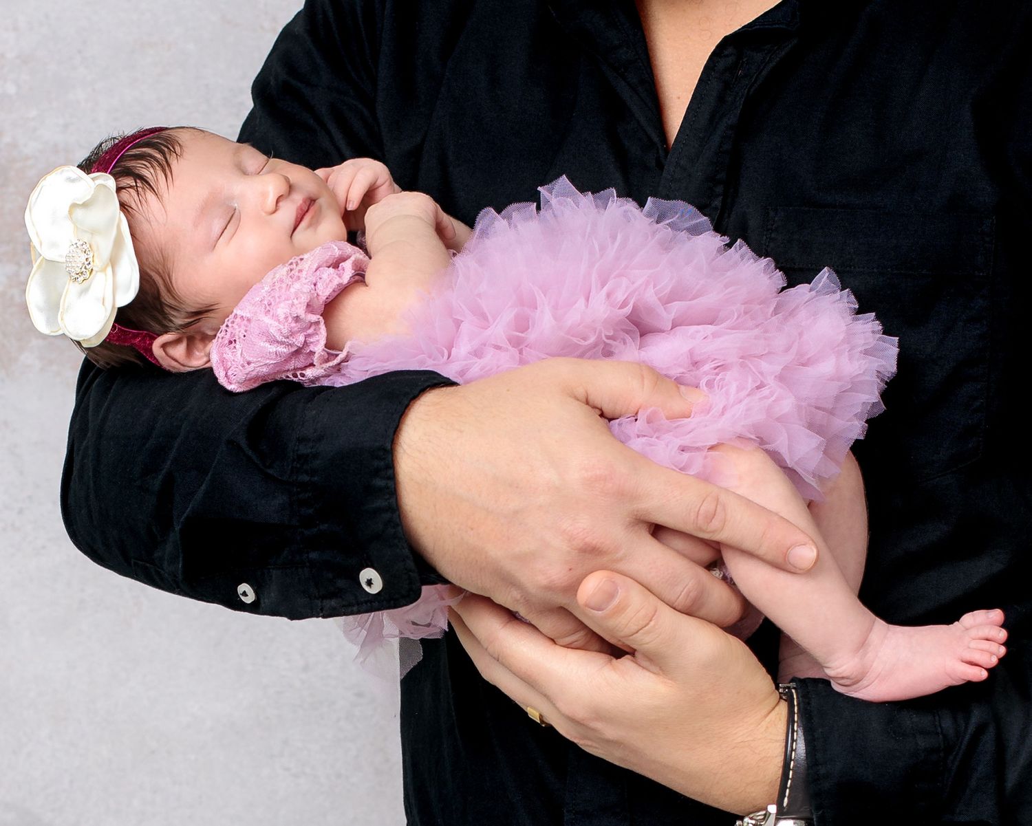 Newborn baby in mauve fluffy dress and flower head band sleeping embraced in her fathers arms taken by Family Photographer Caz Captures.