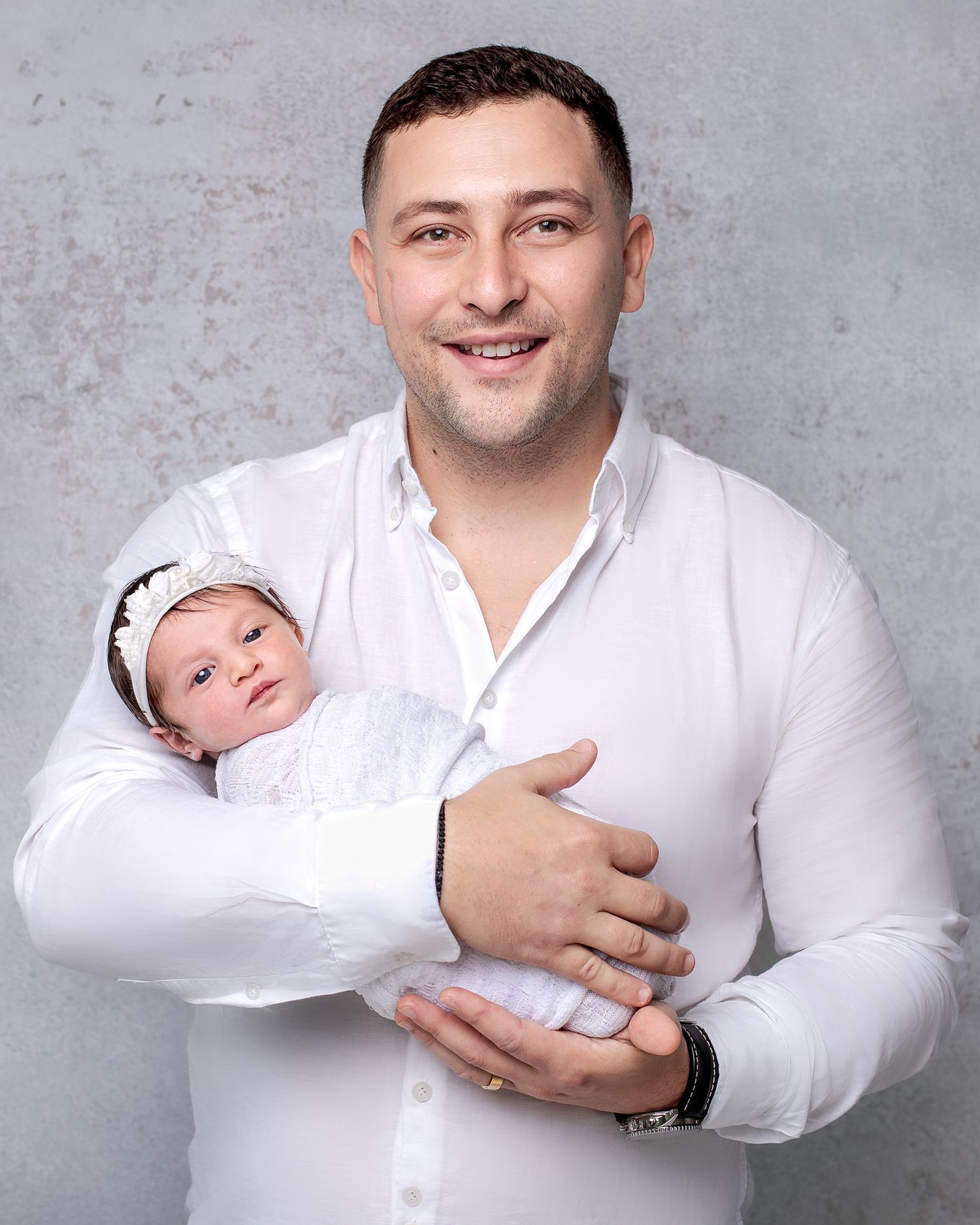 Sydney newborn photoshoot: father in a white shirt holds newborn baby daughter wrapped in white, modern minimalist style.