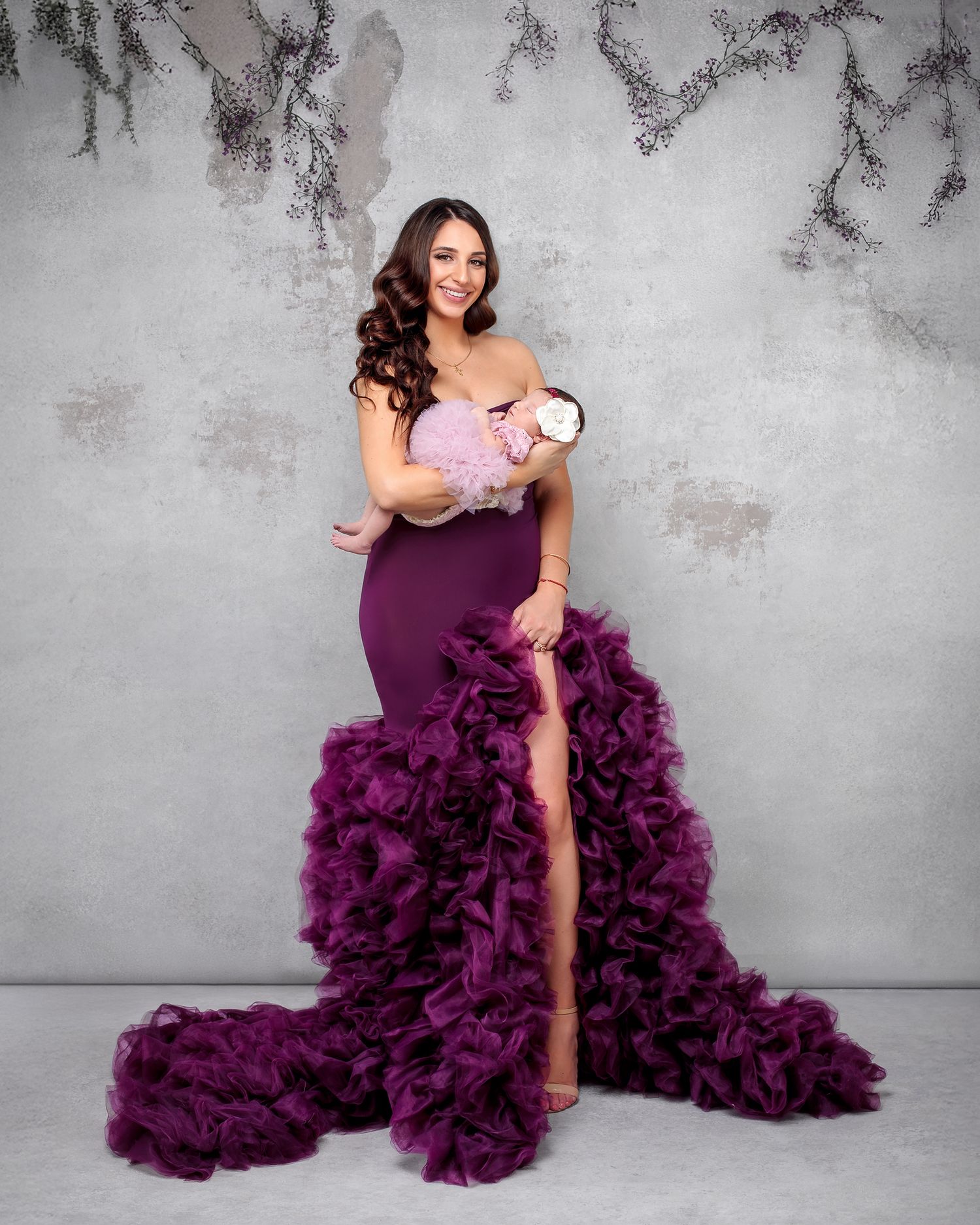 Mother embracing newborn baby girl looking stunning in couture fluffy deep magenta gown, newborn daughter in matching mauve dress.