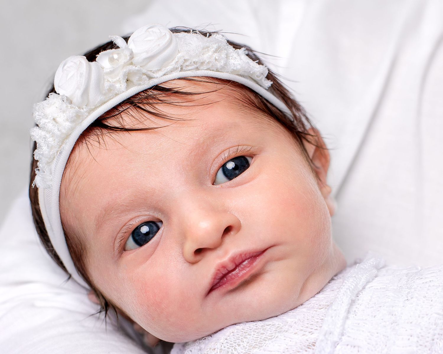 Close-up of baby girls head, as she is held and wrapped in white with white flower headband.