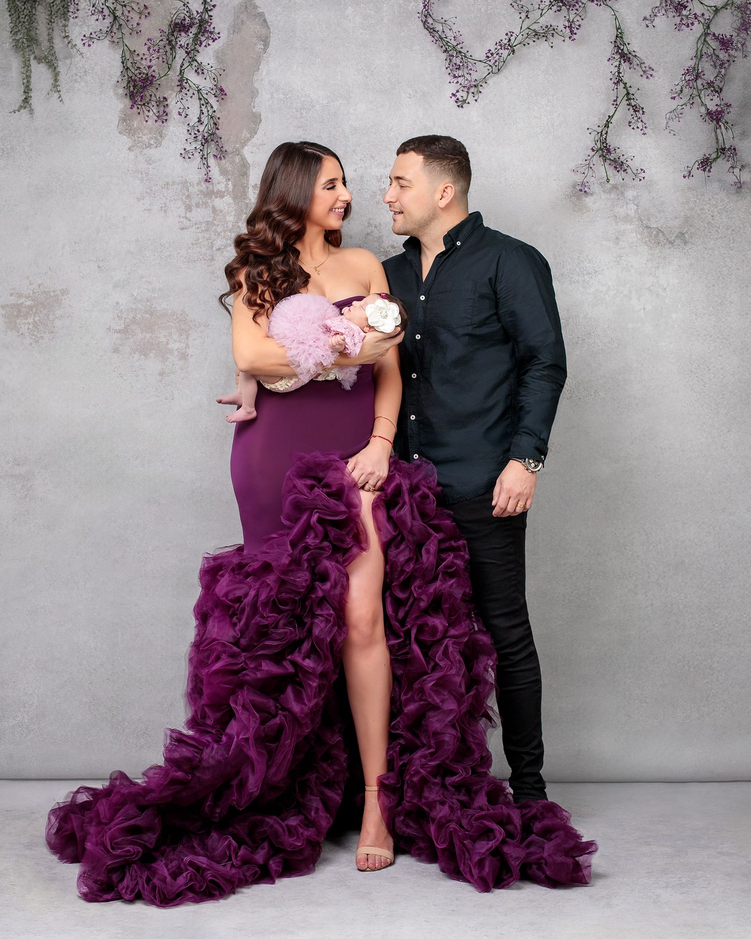 Photo of a glamorous couple smiling holding baby girl , mother wearing fluffy deep purple gown and newborn baby girl in a mauve fluffy dress