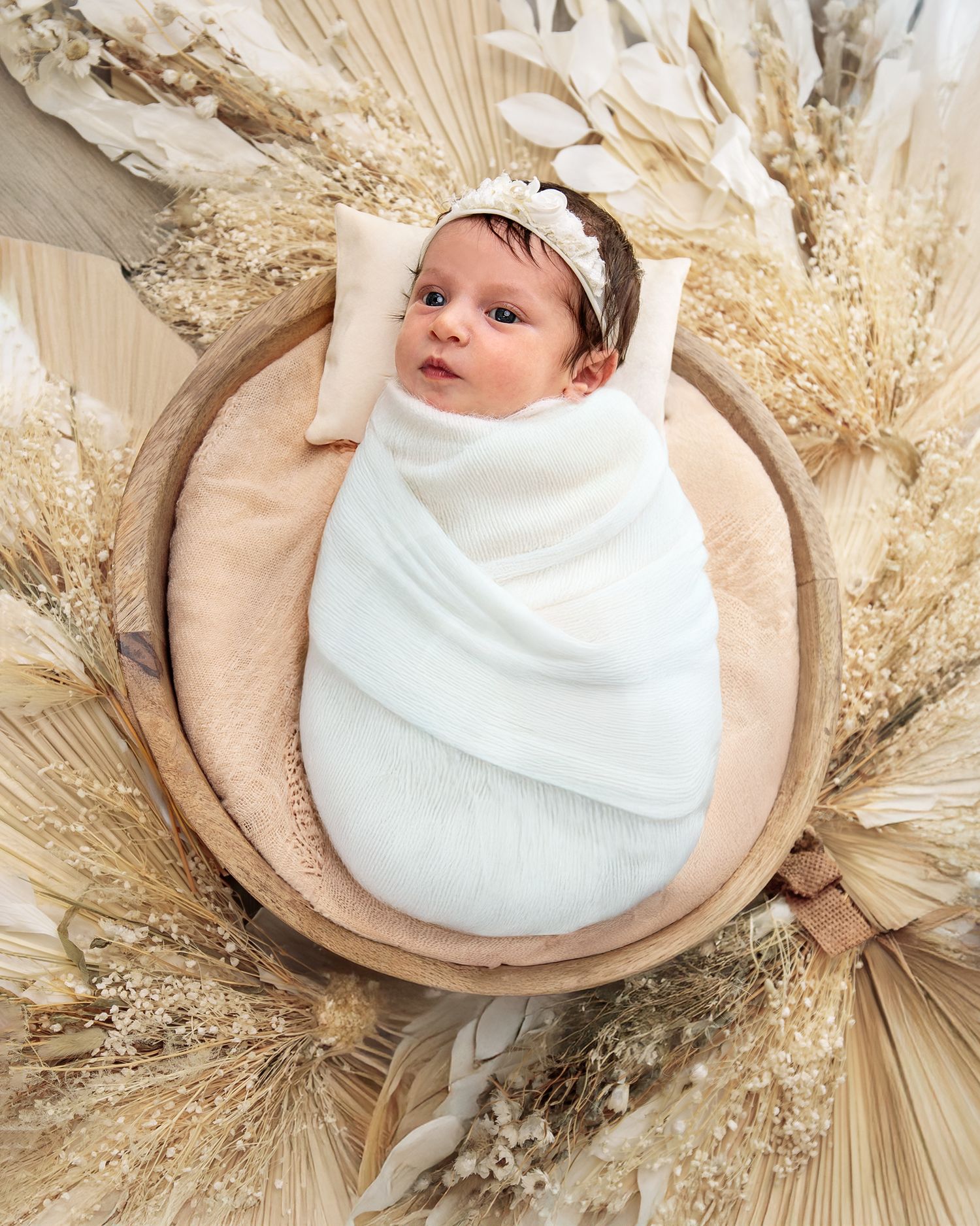 Newborn portrait of baby girl wrapped in white blanket and with matching white headband, wood dough bowl, against dried flower arrangement.