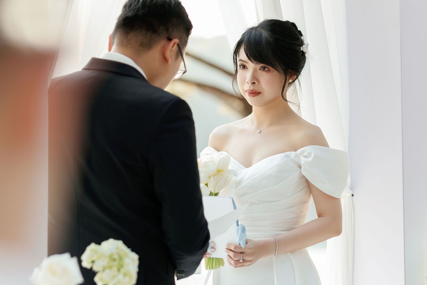 Wedding ceremony with couple in traditional white dress and black suit against window lighting.